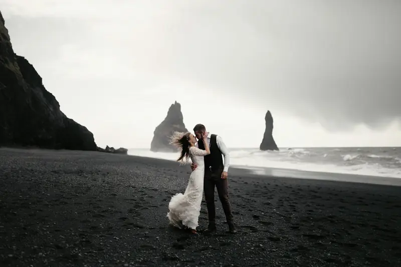 Couple embracing on a dark-sand beach below sea stacks
